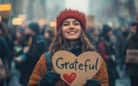 The image shows a young woman holding a heart-shaped sign that reads "Grateful" with a red heart in the center. She is wearing a red knitted beanie and a yellow sweater, and is smiling at the camera. The background is blurred, but it appears to be a busy street with people walking and holding signs. The overall mood of the image is cheerful and positive.
